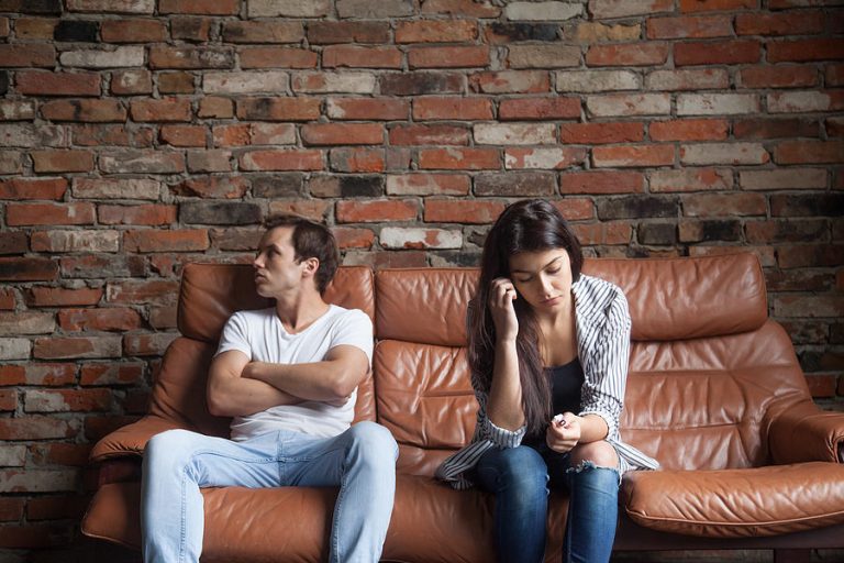 A man and his wife are sitting on a couch.  Man is giving his wife the silent treatment.