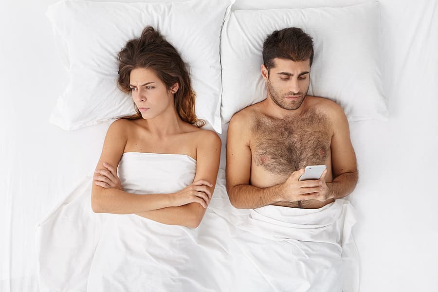 man and wife in bed while man is on his cell phone